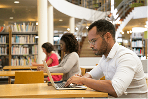 Three people sit at separate tables in a library, working on laptops. Bookshelves and a spiral staircase are visible in the background. The setting is quiet and focused.
