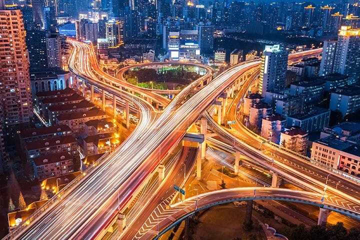 Aerial view of a brightly lit city at night, showing a complex network of elevated highways with streaks of car lights weaving through tall buildings and urban structures.