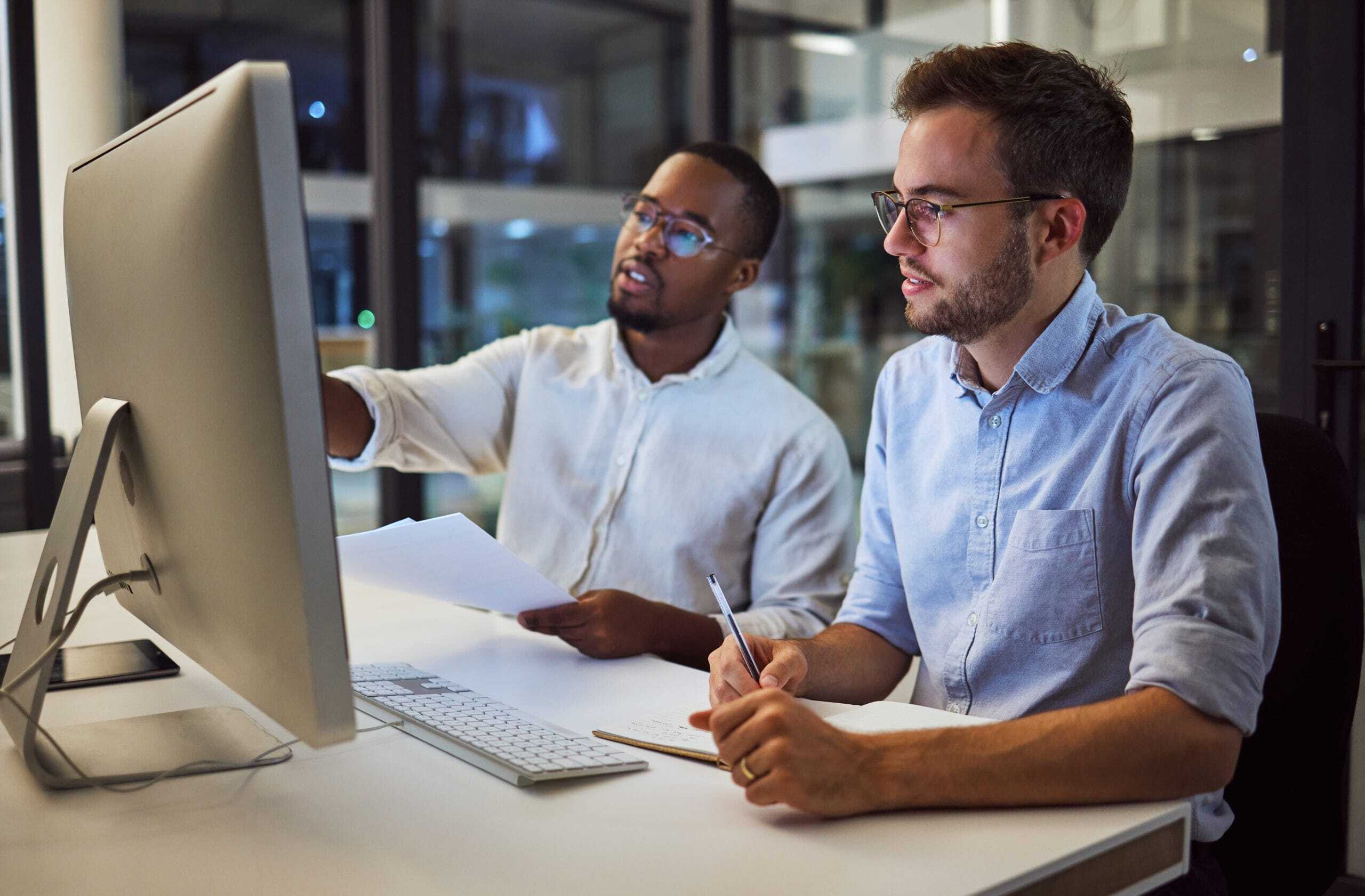 Two men sit at a desk in an office, looking at a computer monitor. One man points at the screen while holding papers; the other man holds a pencil and notebook. Both appear focused and engaged in discussion.