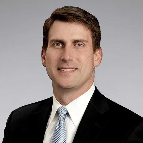 A man in a dark suit, white dress shirt, and light blue patterned tie, smiling against a plain gray background.