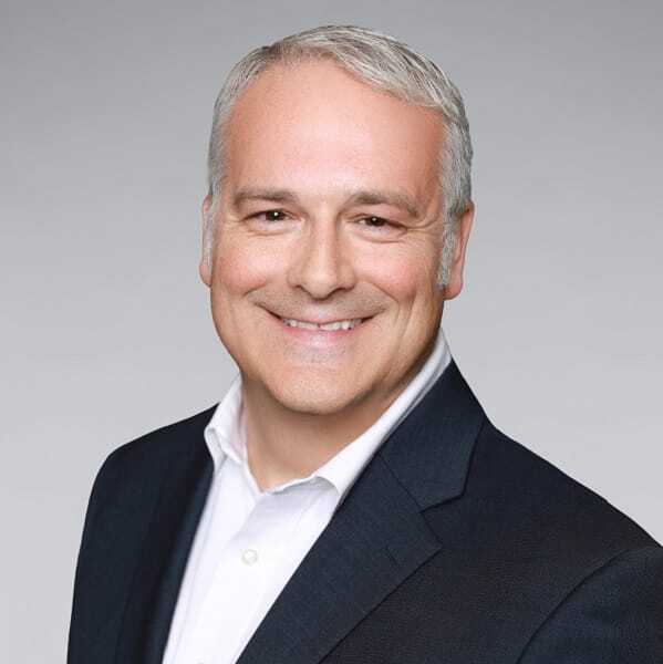 A smiling middle-aged man with short gray hair, wearing a dark suit jacket and a white collared shirt, poses in front of a light gray background.