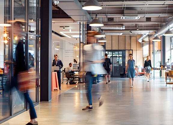 Modern office space with people walking and working; glass walls, open seating, and industrial-style lighting create a contemporary, collaborative environment. Some people are in motion while others sit at desks.