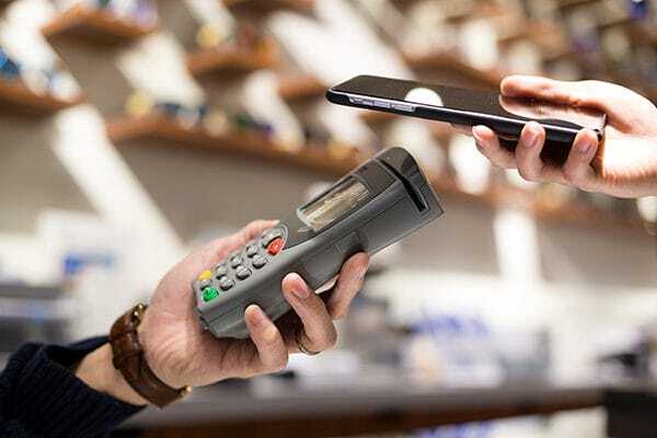A person holds a payment terminal while another person pays using a smartphone, indicating a contactless mobile payment in a store setting.