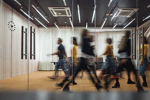Several people walk briskly through a modern office hallway with glass walls and a visible clock on the wall; their motion appears blurred.