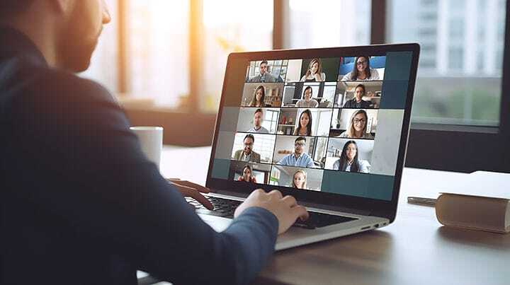 A person sits at a desk using a laptop for a video conference, with multiple people visible in small windows on the screen. Sunlight streams through large windows in the background.